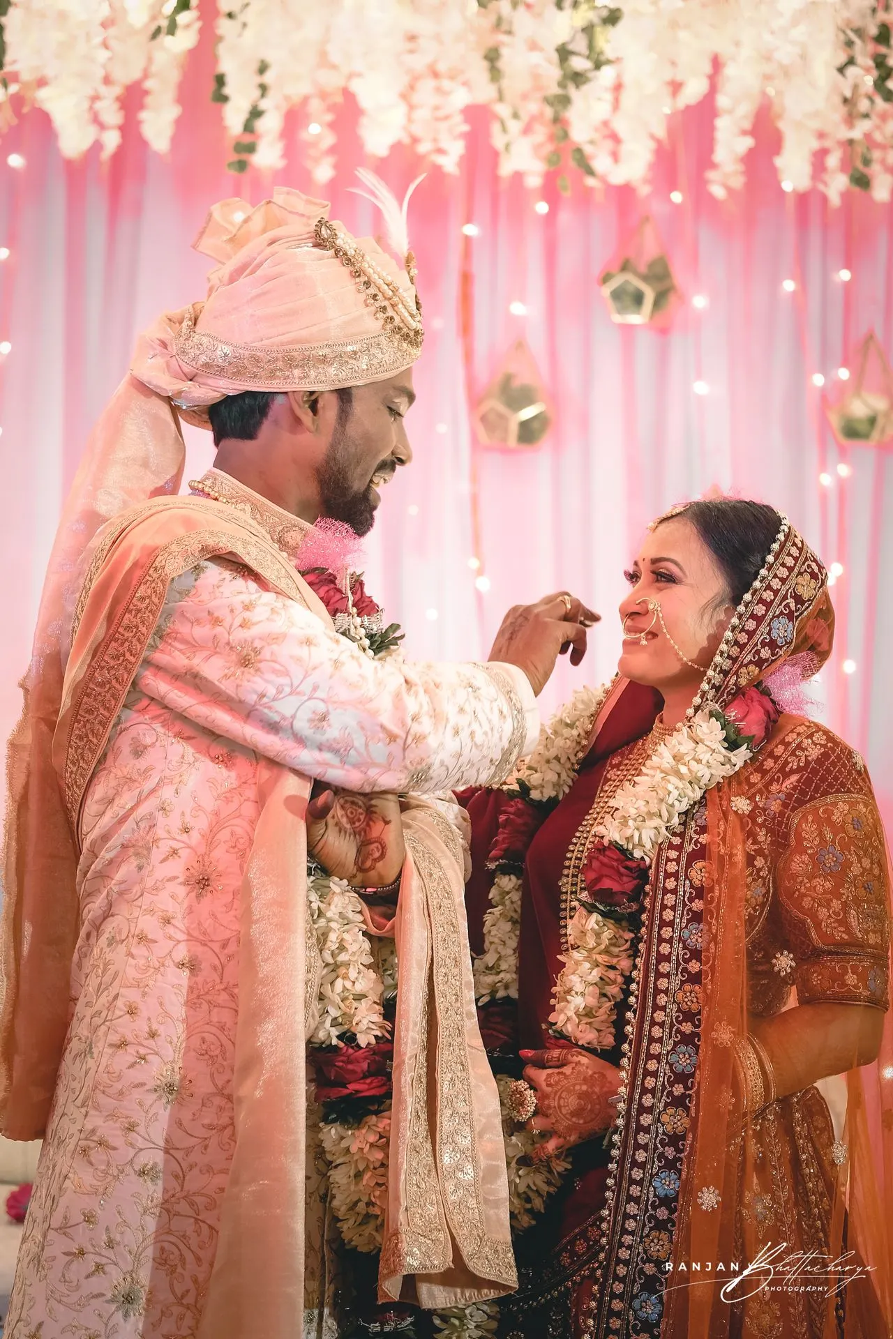Ruhi & Roshan’s Wedding Ceremony - Captured by Ranjan Bhattacharya in Bihar A wedding photograph featuring the bride, Ruhi Singh, and the groom, Roshan Kumar, in traditional Indian attire during a ceremony. The bride is adorned in an ornate orange saree with intricate embroidery, while the groom is dressed in a cream sherwani with pink detailing. They are standing under a floral canopy with white flowers and strings of lights, creating a festive and elegant atmosphere. The image captures a moment where the groom appears to be placing a garland around the bride’s neck, symbolizing their union. Photography by Ranjan Bhattacharya, captured in Bihar.