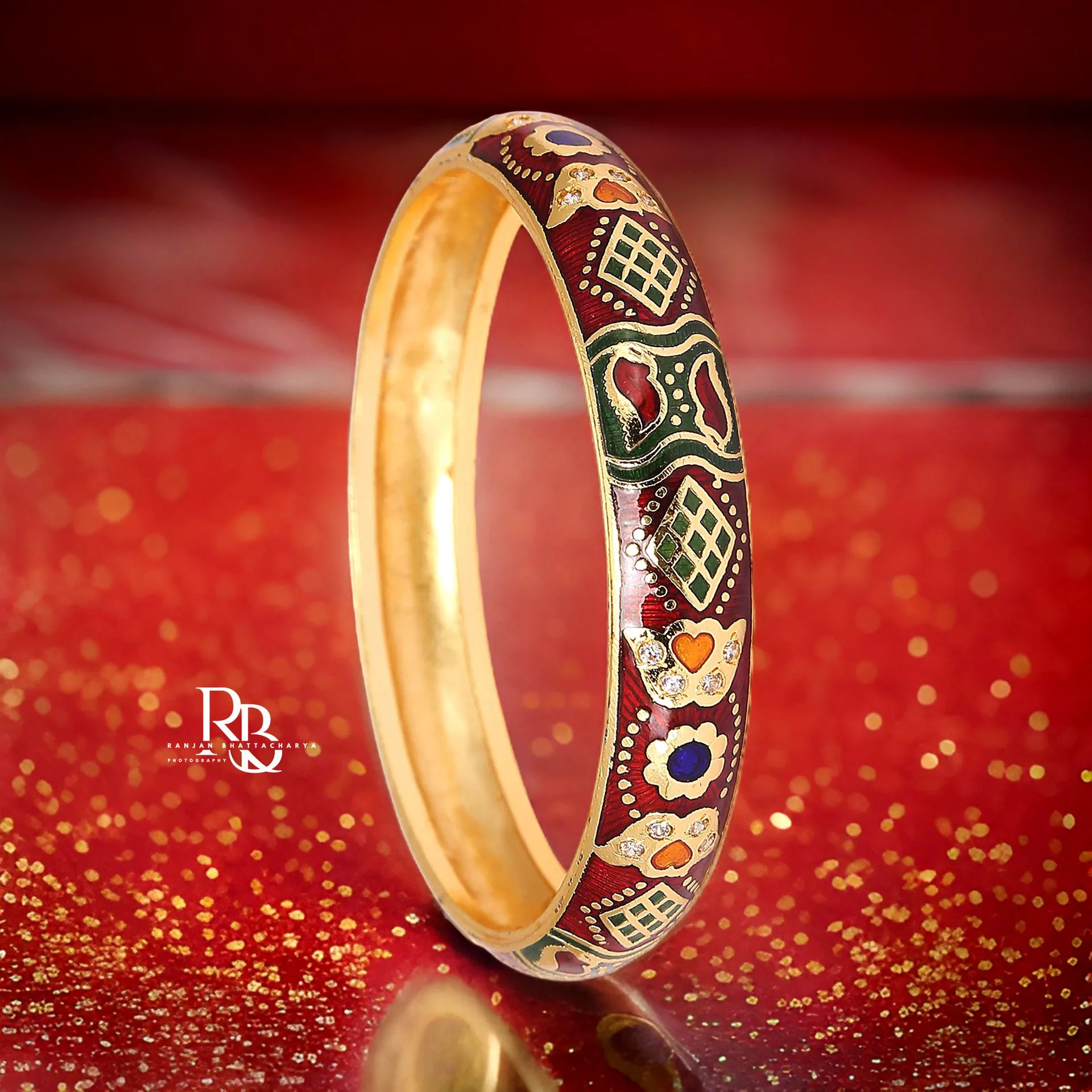 A traditional gold bangle with detailed enamel work in red, green, and gold colors, embellished with intricate patterns and hearts, photographed by Ranjan Bhattacharya.