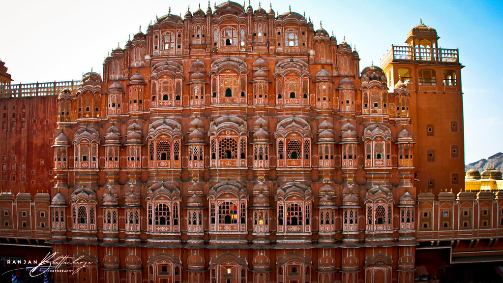 The façade of Hawa Mahal, a grand and ornate five-story building made of red and pink sandstone, featuring an intricate pattern of small windows and carved balconies. Photo by Ranjan Bhattacharya.