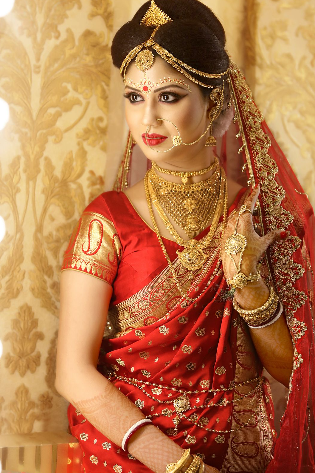 Bride Dr. Debarati Sarkar in traditional bridal attire and jewelry, getting final touches on her hairstyle by hands adorned with red bangles, against a softly blurred warm-lit background.