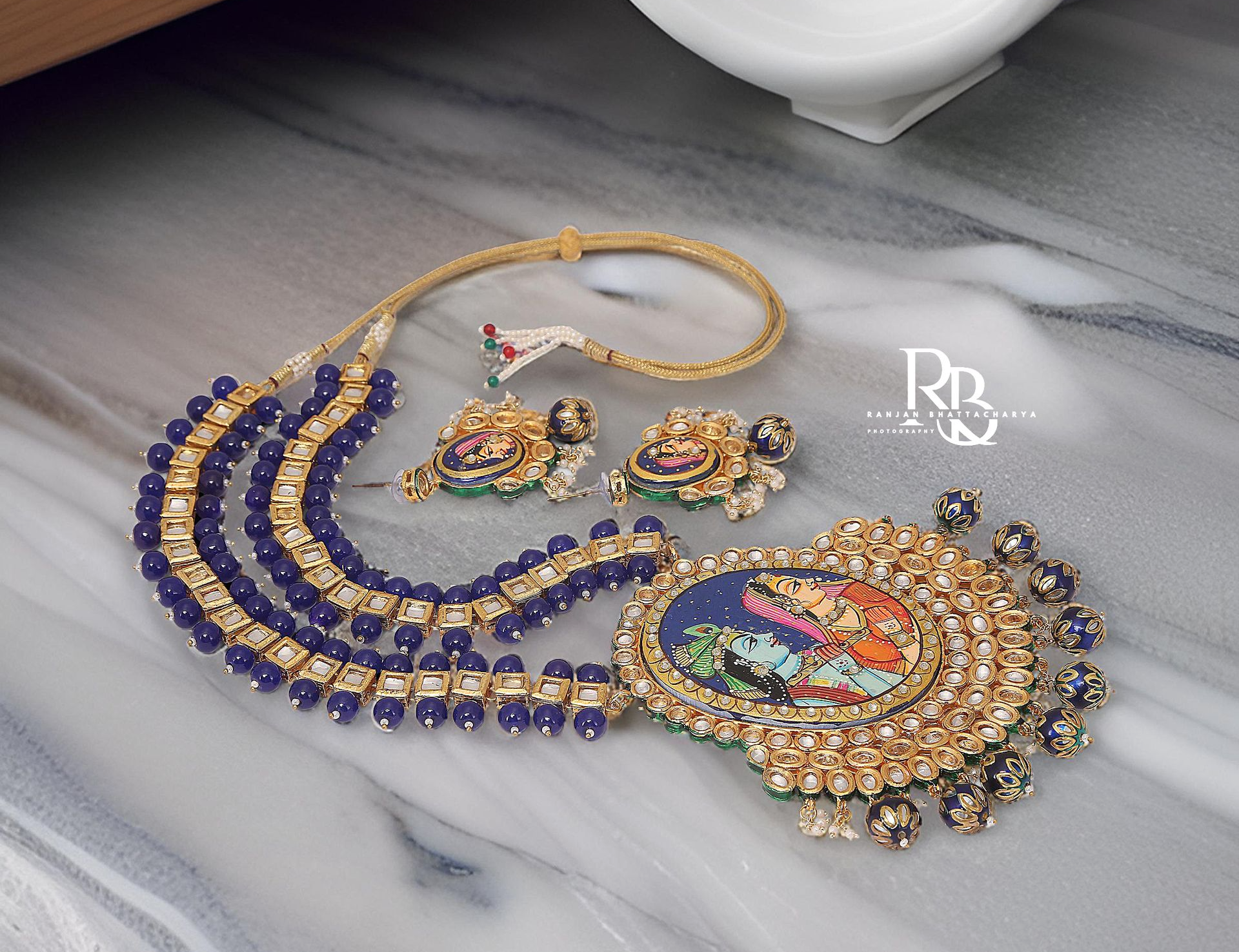 Handcrafted necklace and earrings set with Krishna & Radha Pichwai art and blue bead embellishments on a table, photographed by Ranjan Bhattacharya.